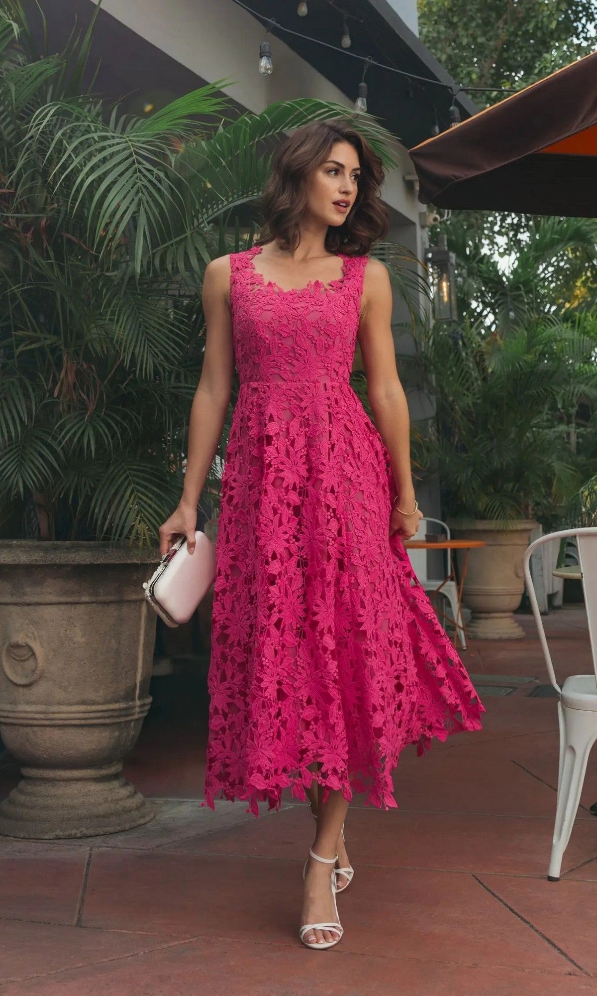 A woman in the hot pink Mac Duggal 69073 midi party dress, sleeveless and floral lace, holding a clutch while standing outdoors near white chairs and green plants.